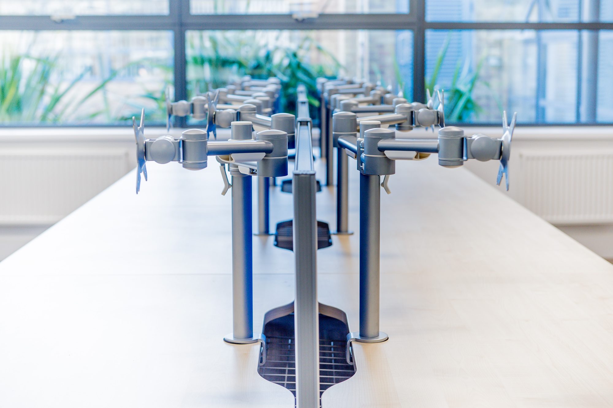 Row of monitor arms on bench desks, looking out to the trees
