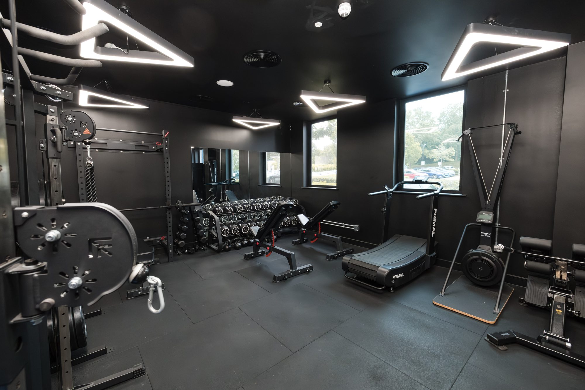 On-site gym room with dark walls, triangular feature lighting, free weights, treadmill and resistance machines
