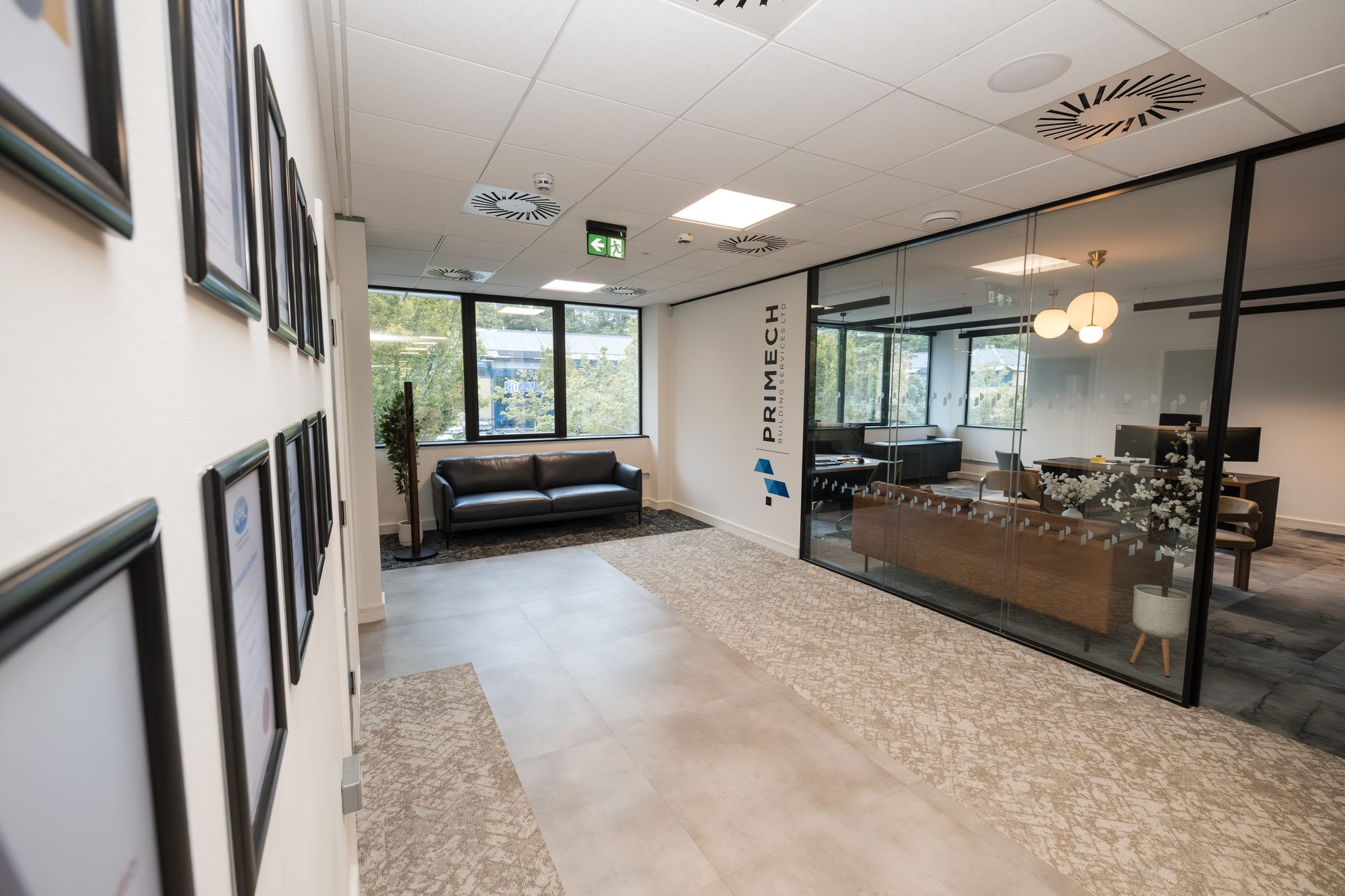 Primech reception with PRIMECH branding wall visible through glazed partition, leather sofa and framed certifications on the wall